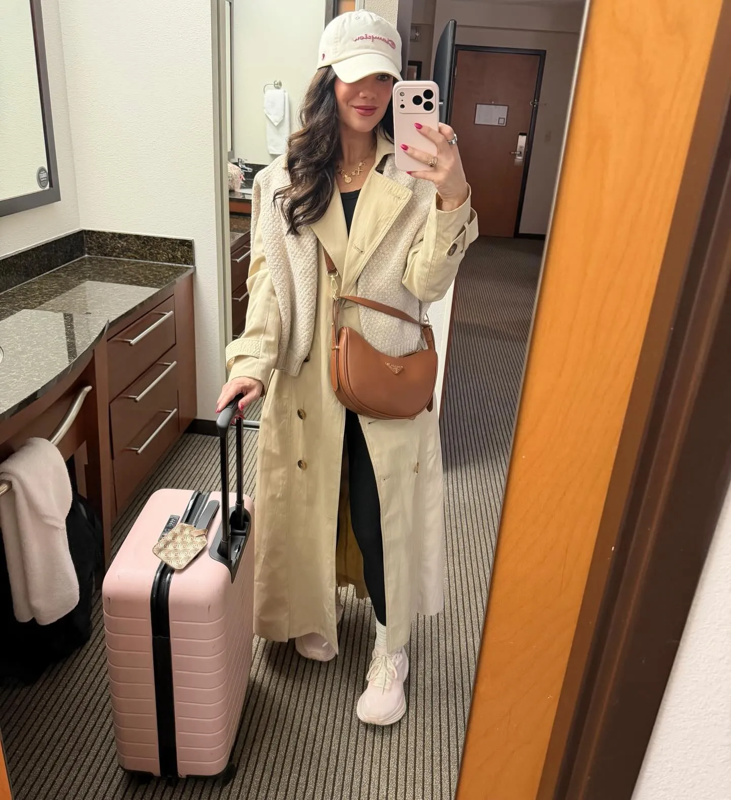 Susanne in a cream ball cap and trench coat with black tank and leggings, tan crossbody bag, blush sneakers and a pink carry-on suitcase