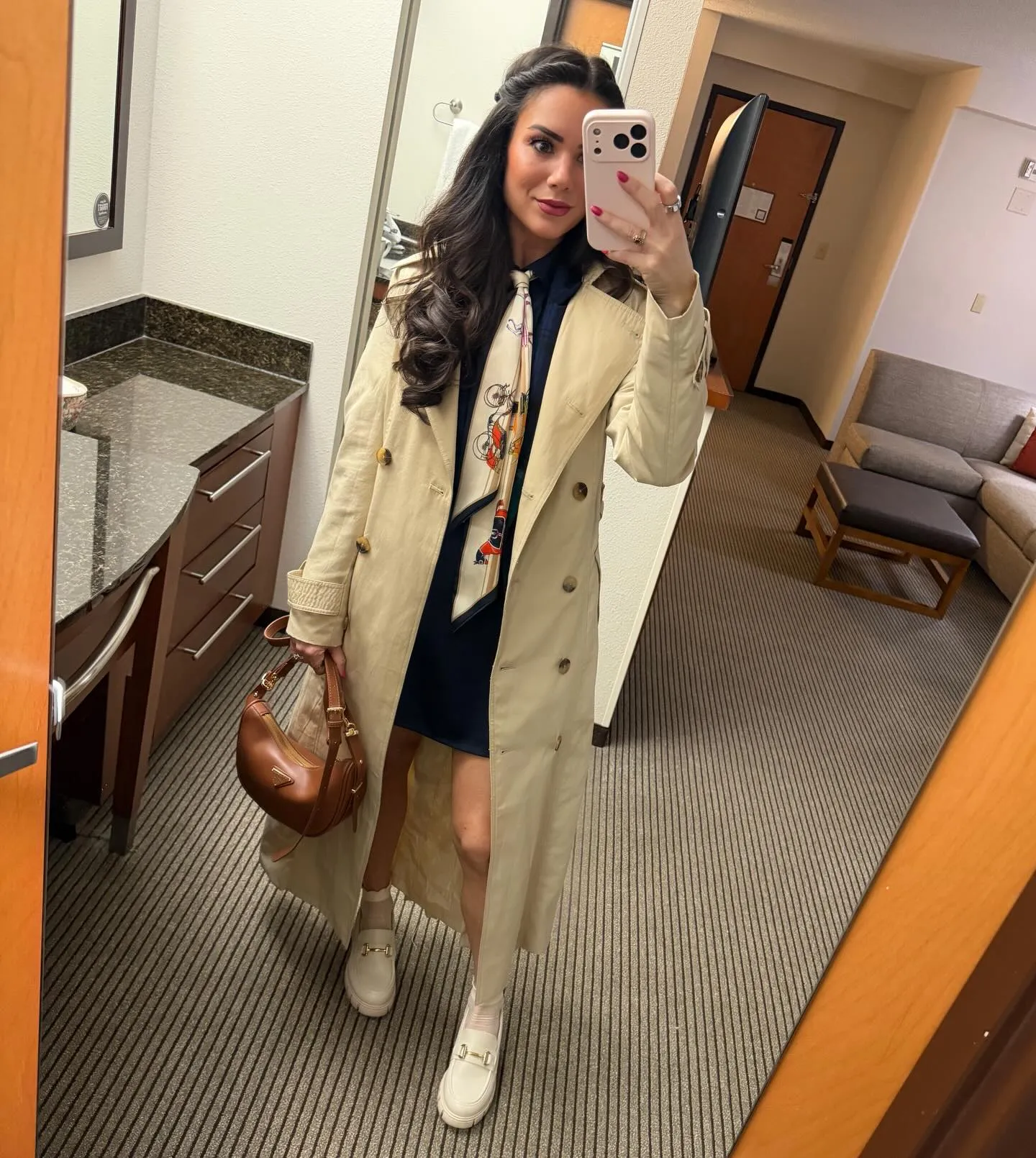 Susanne in a long tan trench coat over a navy dress with a cream printed scarf tie, brown shoulder bag and cream loafers with sheer socks
