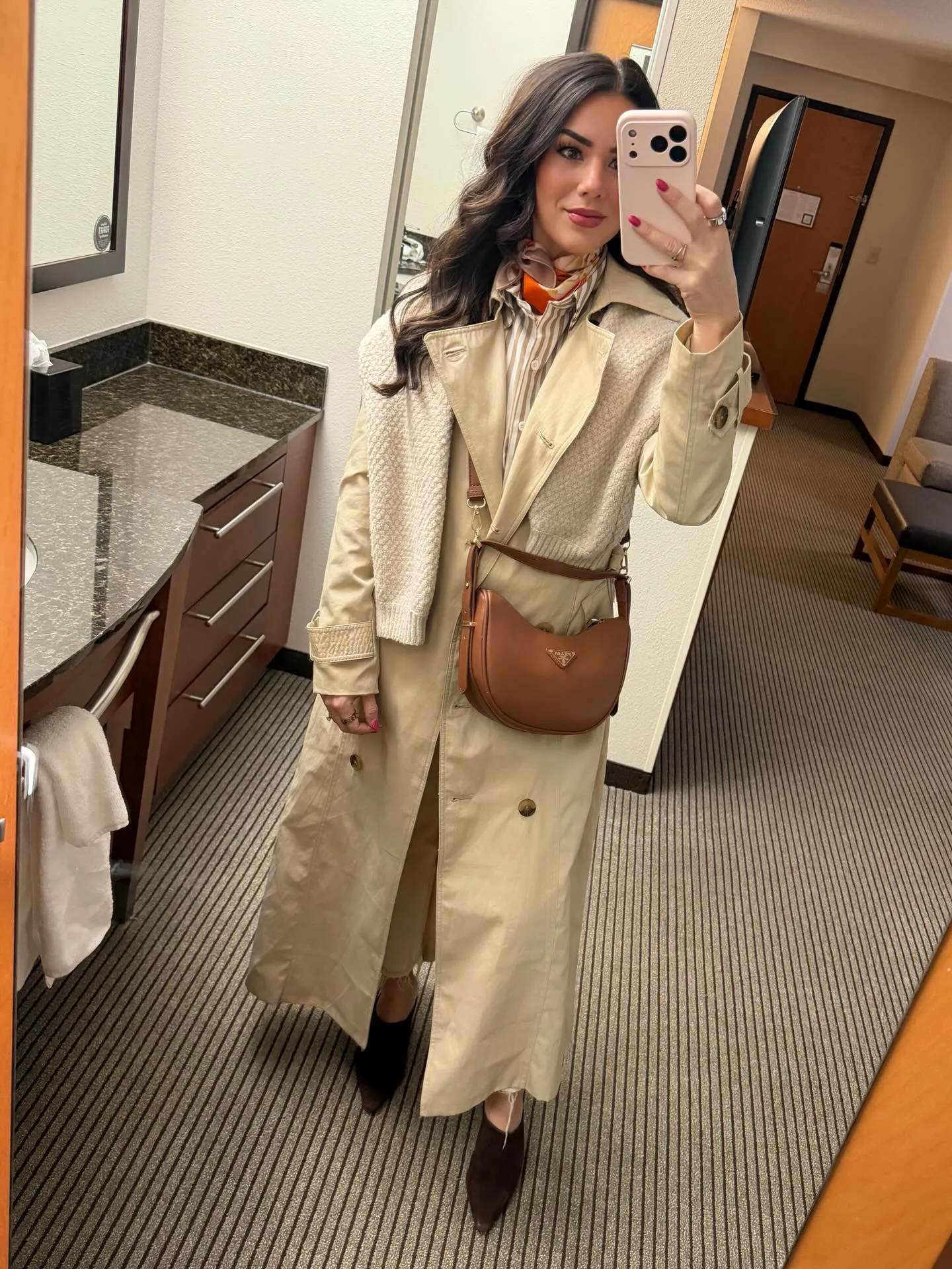 Susanne in a cream trench coat with a knit sweater over one shoulder, orange scarf at her neck and a brown crossbody bag in a hotel room mirror