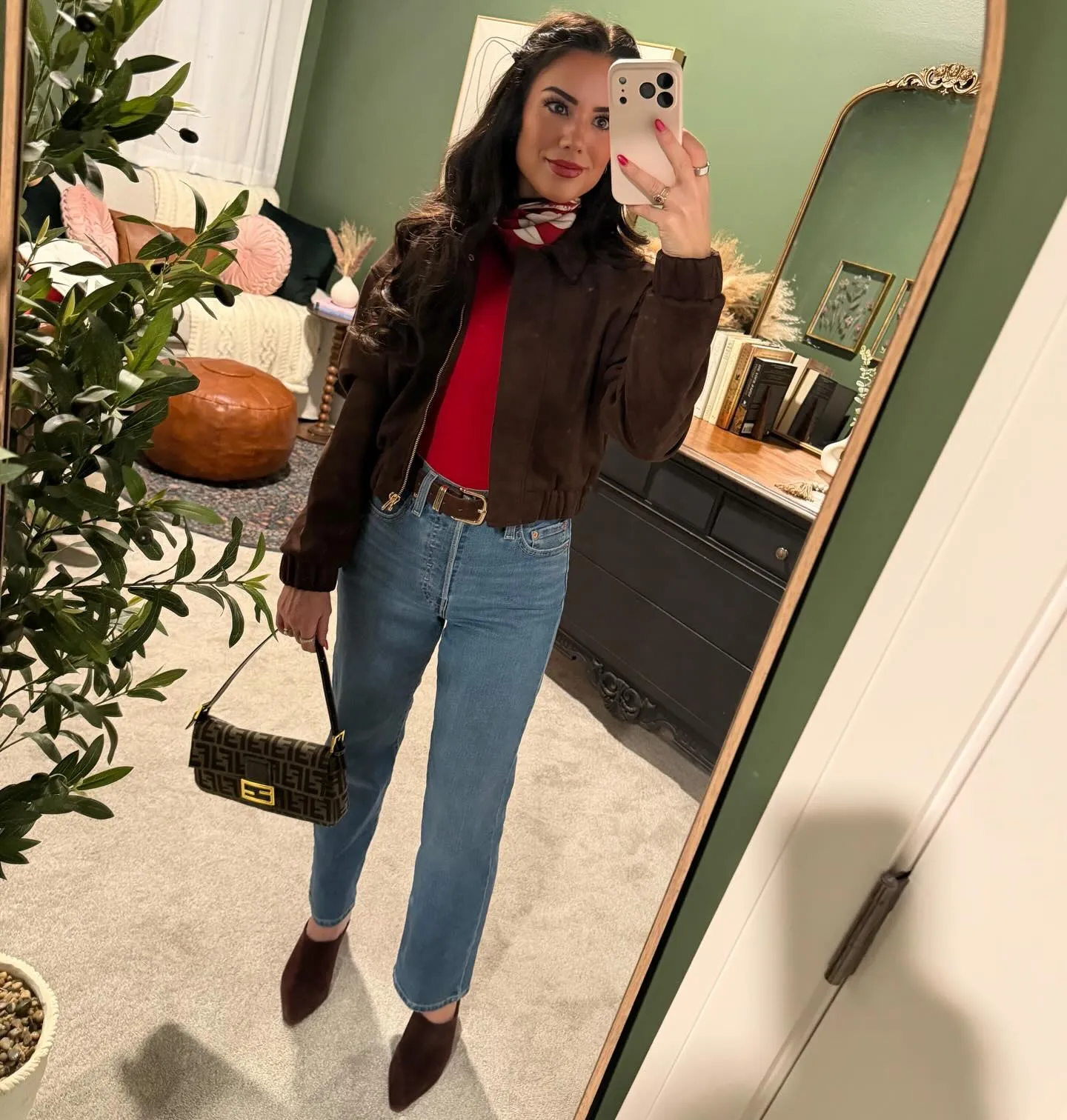 Susanne in a brown suede bomber over a red top with a red and white scarf at her neck, medium-wash jeans, brown baguette bag and chocolate suede mules