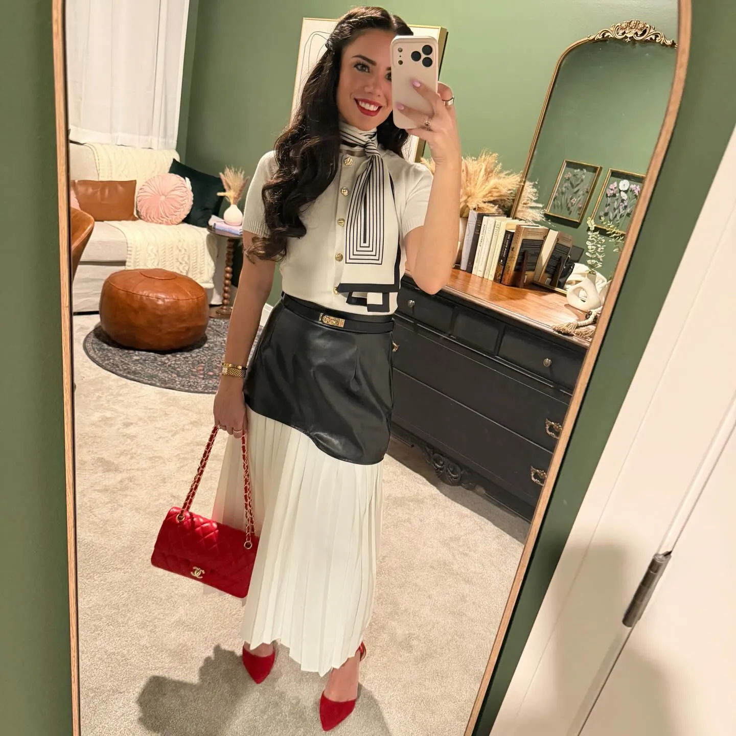 Susanne in a white sweater with gold buttons, striped scarf tied like a tie, black leather and white pleated maxi skirt, red quilted bag and red pumps