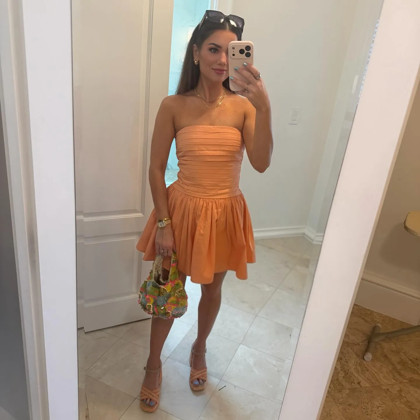 Susanne in a strapless orange mini dress with a pleated bodice and flared skirt, colorful beaded bag and orange raffia platforms in a hotel mirror