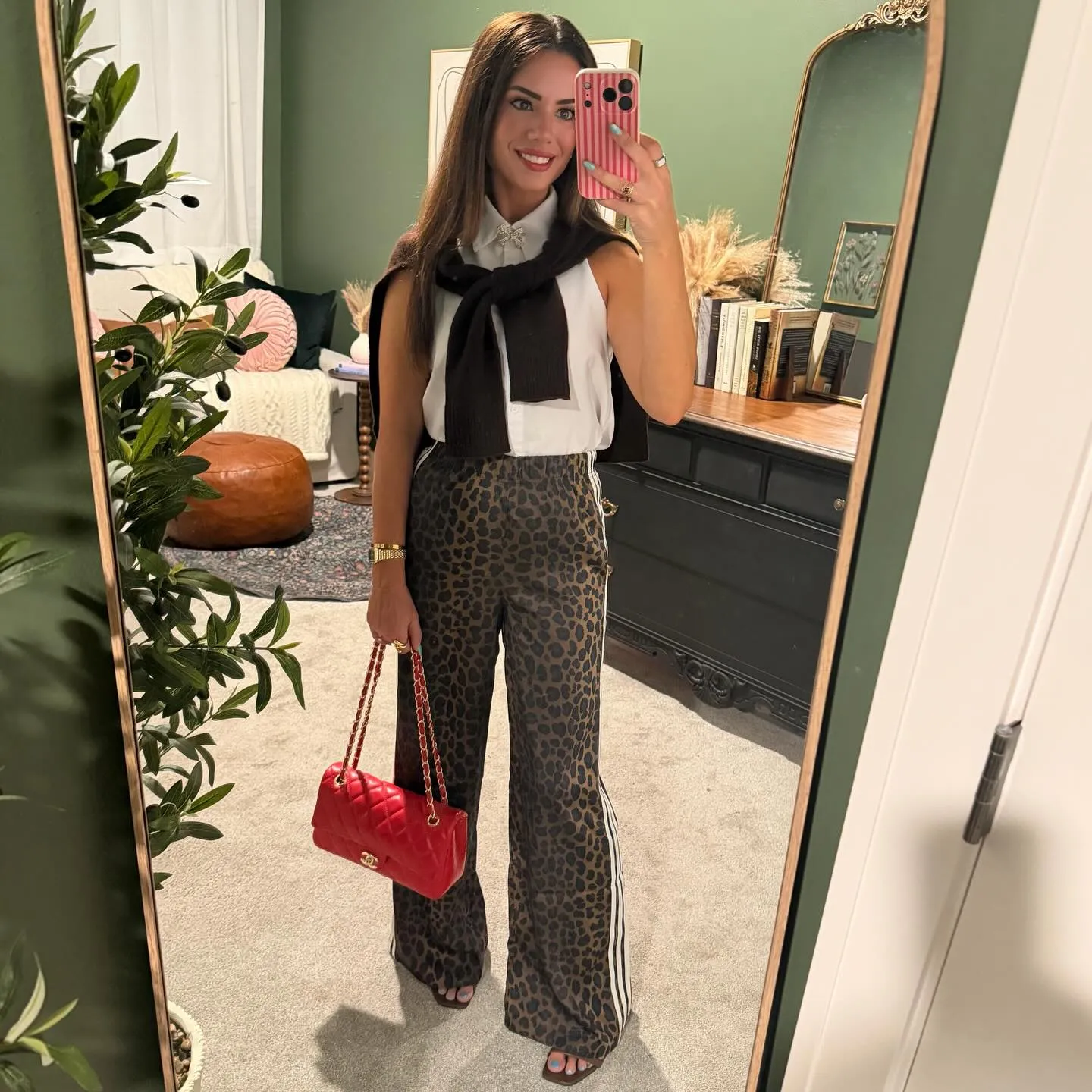 Susanne in a white sleeveless button-down with a bow brooch, brown leopard track pants, dark sweater tied over her shoulders and a red quilted bag