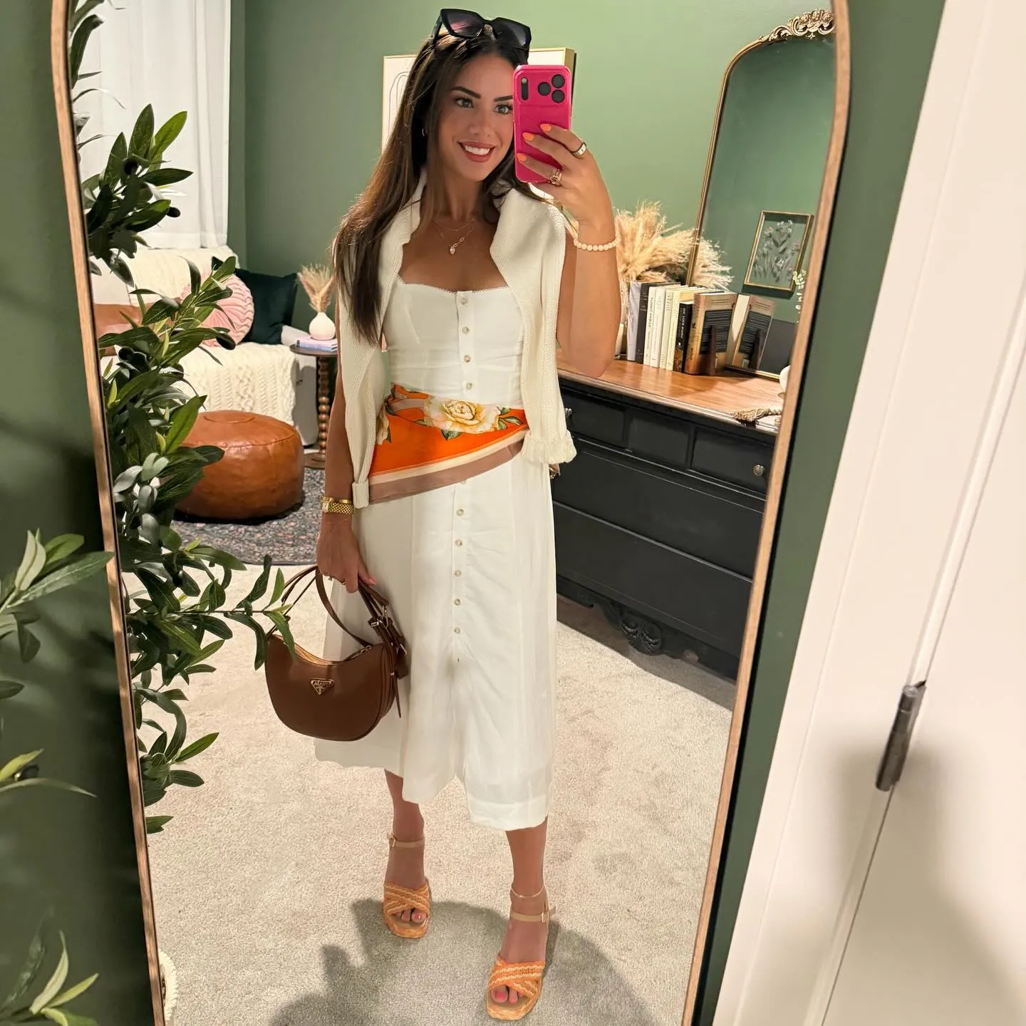Susanne in a white button-front sundress with an orange floral waist scarf and cream sweater over her shoulders, mirror selfie in her green dressing room
