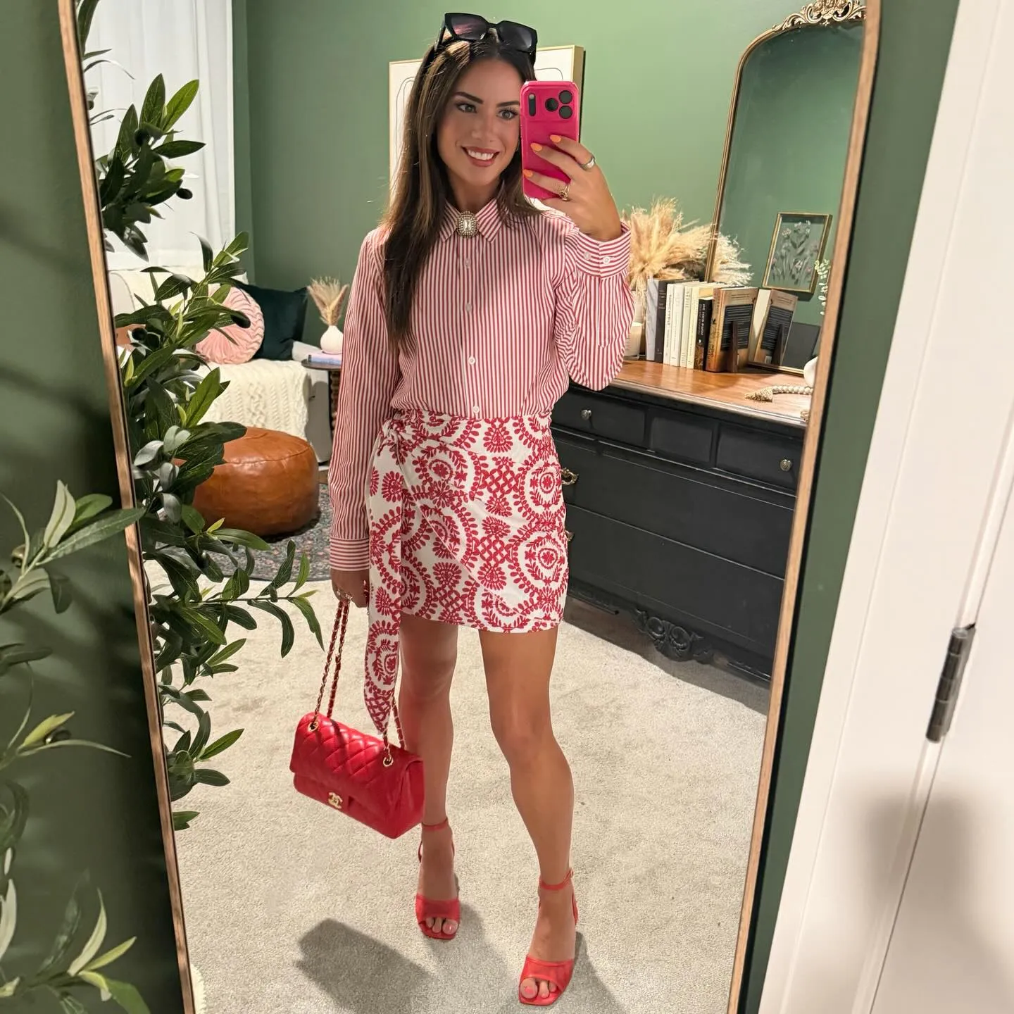 Susanne in a red and white striped button-down with a gold brooch at the collar, a red and white floral wrap-front mini skirt, red heels, and a red quilted chain bag, mirror selfie in her green dressing room