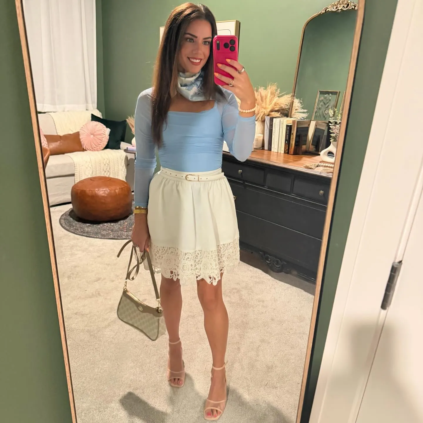 Mirror selfie in a light blue long-sleeve mesh bodysuit with a blue floral neck scarf, white lace-trim mini skirt, belt, tan bag and nude heels.